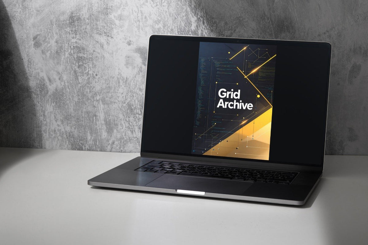 A silver laptop sits open on a white surface against a textured gray wall, displaying a digital cover titled 'Grid Archive' on its screen.